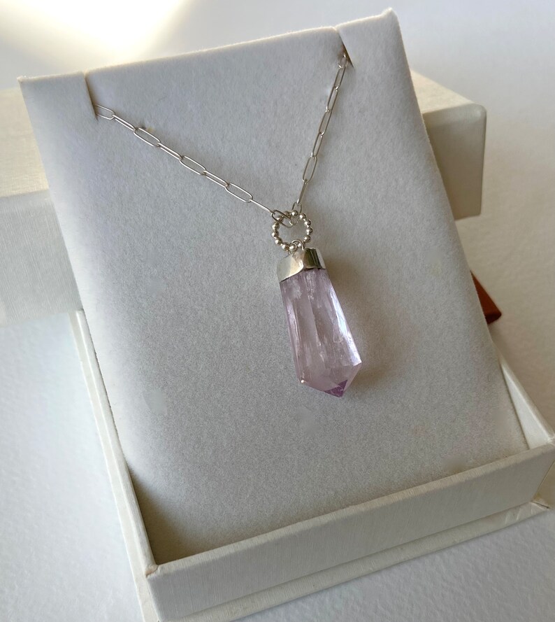 Puede incluir: Un collar de cadena de plata con un colgante de cristal rosa p&aacute;lido en una caja blanca. El cristal est&aacute; facetado y tiene una punta puntiaguda.