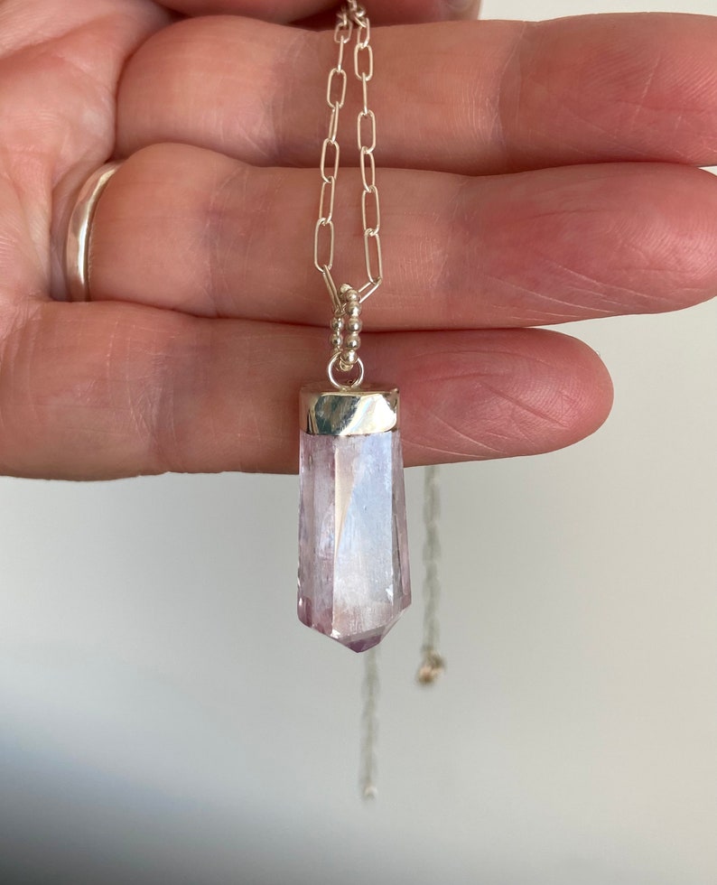 Puede incluir: Un collar de cadena de plata con un colgante de cristal transparente, facetado, de color p&uacute;rpura claro.