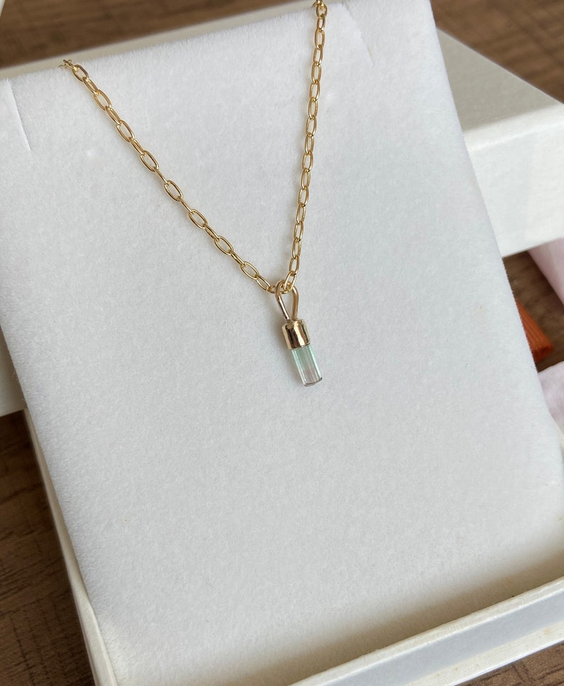 May include: A gold chain necklace with a light blue crystal pendant. The pendant is a long, thin rectangle.
