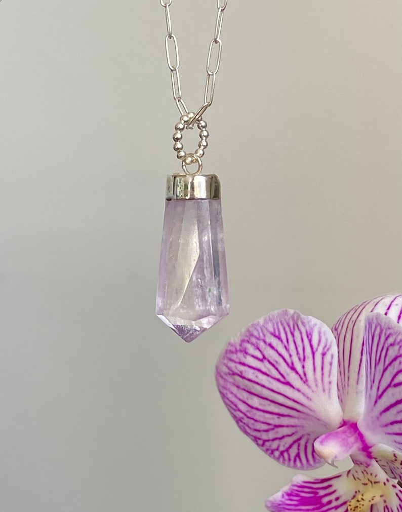 Puede incluir: Un collar de cadena de plata con un colgante de cristal morado claro en forma de punta. El colgante est&aacute; engastado en un bisel de plata.