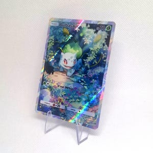 Könnte beinhalten: Eine holografische Bulbasaur-Sammelkarte mit einem blauen Wesen mit einer grünen Zwiebel auf dem Rücken in einer Waldszene. Die Karte zeigt den Text Bulbasaur, HP 70 und Vine Whip 40, mit einer regenbogenfarbenen Lichtreflexion auf der Oberfläche.