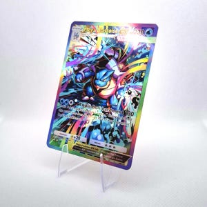 Puede incluir: Una carta coleccionable holográfica de Mega Blastoise EX que muestra una criatura azul similar a una tortuga con cañones en la espalda. La carta presenta 230 HP, el ataque Triple Bombardment y rayas de luz coloridas. Se sostiene en posición vertical mediante un soporte de plástico transparente.