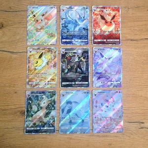 May include: A collection of nine holographic trading cards featuring Eevee and its evolutions, including Vaporeon, Flareon, Jolteon, Umbreon, Espeon, Leafeon, Glaceon, and Sylveon. Each card displays colorful character illustrations, text, and game stats.