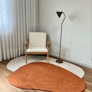 Puede incluir: Un interior moderno con un sill&oacute;n crema con brazos de madera. Una alfombra en forma org&aacute;nica de color &oacute;xido se encuentra sobre una alfombra beige claro. Una l&aacute;mpara de pie negra y cortinas transparentes completan la escena.