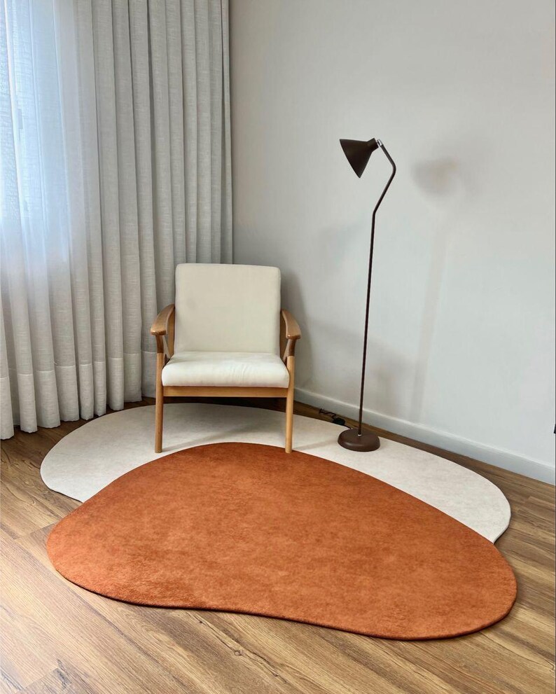 Puede incluir: Un interior moderno con un sill&oacute;n crema con brazos de madera. Una alfombra en forma org&aacute;nica de color &oacute;xido se encuentra sobre una alfombra beige claro. Una l&aacute;mpara de pie negra y cortinas transparentes completan la escena.