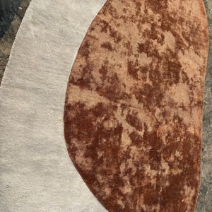 Puede incluir: Una alfombra abstracta con una secci&oacute;n ovalada gris claro y una secci&oacute;n m&aacute;s grande, marr&oacute;n, en forma de ri&ntilde;&oacute;n con una textura moteada. El dise&ntilde;o de la alfombra crea una est&eacute;tica &uacute;nica y contempor&aacute;nea, adecuada para varios interiores.