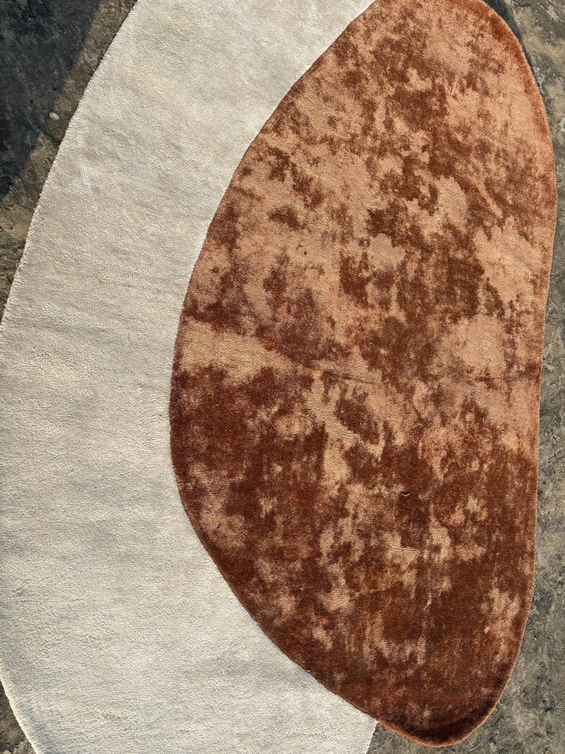 Puede incluir: Una alfombra abstracta con una secci&oacute;n ovalada gris claro y una secci&oacute;n m&aacute;s grande, marr&oacute;n, en forma de ri&ntilde;&oacute;n con una textura moteada. El dise&ntilde;o de la alfombra crea una est&eacute;tica &uacute;nica y contempor&aacute;nea, adecuada para varios interiores.
