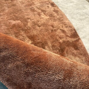 Puede incluir: Primer plano de una alfombra enrollada, afelpada, de color &oacute;xido, con una superficie suave y texturizada. La alfombra tiene un dise&ntilde;o curvo con una secci&oacute;n beige claro y una secci&oacute;n marr&oacute;n m&aacute;s oscura. La alfombra parece estar hecha de un material suave de pelo alto.