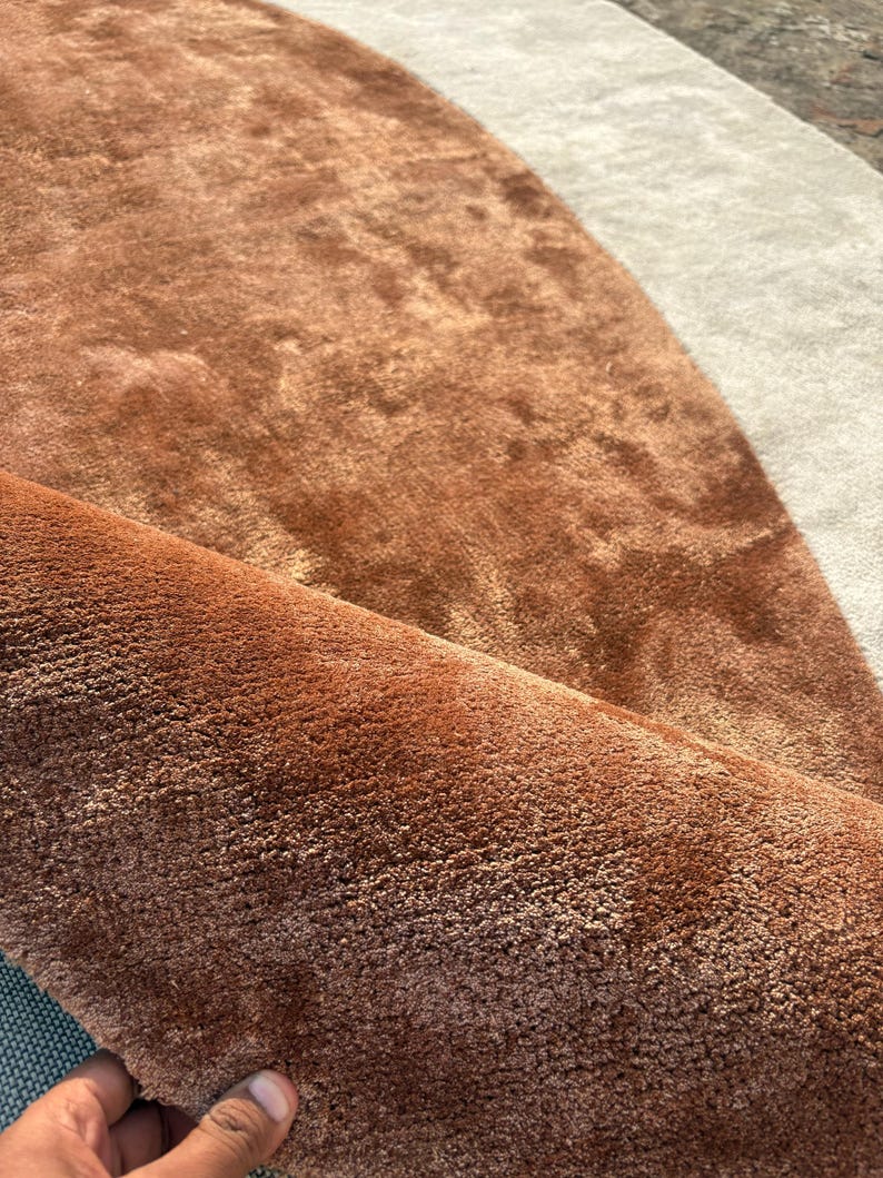 Puede incluir: Primer plano de una alfombra enrollada, afelpada, de color &oacute;xido, con una superficie suave y texturizada. La alfombra tiene un dise&ntilde;o curvo con una secci&oacute;n beige claro y una secci&oacute;n marr&oacute;n m&aacute;s oscura. La alfombra parece estar hecha de un material suave de pelo alto.
