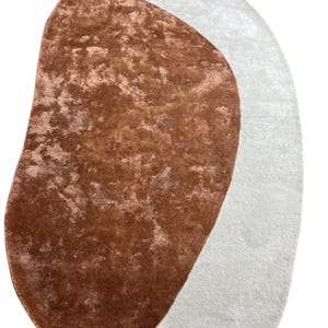 Puede incluir: Dos alfombras abstractas. Una es una alfombra grande, de forma irregular, en un color marr&oacute;n rojizo c&aacute;lido con un aspecto texturizado. La otra alfombra tiene una forma similar, pero en un color blanco roto suave. Ambas alfombras tienen una textura afelpada y acogedora.