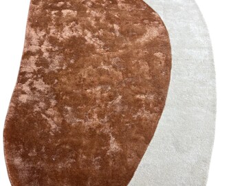 Könnte beinhalten: Zwei abstrakte Teppiche. Einer ist ein großer, unregelmäßig geformter Teppich in einem warmen, rötlich-braunen Farbton mit strukturiertem Aussehen. Der andere Teppich hat eine ähnliche Form, aber in einem weichen, cremefarbenen Farbton. Beide Teppiche haben eine weiche, einladende Textur.