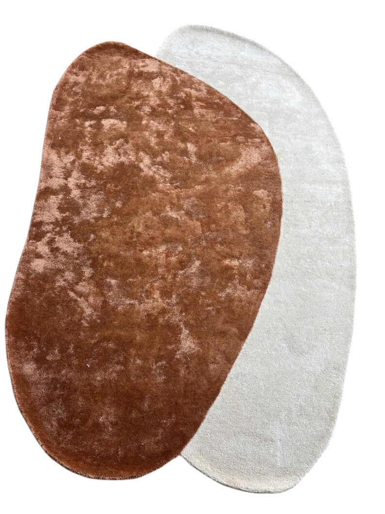 Puede incluir: Dos alfombras abstractas. Una es una alfombra grande, de forma irregular, en un color marr&oacute;n rojizo c&aacute;lido con un aspecto texturizado. La otra alfombra tiene una forma similar, pero en un color blanco roto suave. Ambas alfombras tienen una textura afelpada y acogedora.