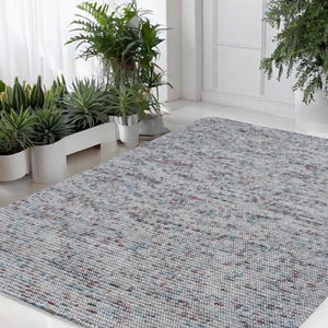 May include: A rectangular area rug with a speckled pattern in shades of gray, blue, and burgundy. The rug is made of a woven material and is placed on a white floor. Several potted plants are in the background.