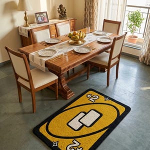 May include: A yellow and black rug in the shape of an Uno card, featuring "+2" text in the corners and two white rectangles. The rug is on a grey tiled floor in a dining room, with a wooden table and chairs.
