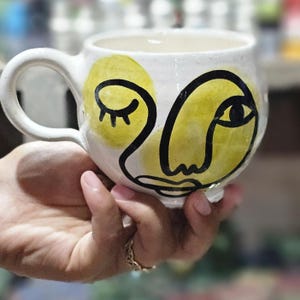 Puede incluir: Taza de cerámica blanca con forma redondeada y asa. La taza presenta un diseño de rostro abstracto amarillo y negro. La cara tiene un ojo cerrado, un ojo abierto, una nariz y una boca. La taza se sostiene en una mano.