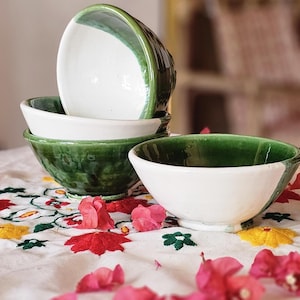 May include: A collection of ceramic bowls, white with a green rim and exterior. The bowls are stacked on a white tablecloth with colourful floral embroidery and pink flowers.