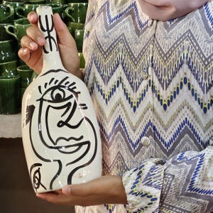 Peut inclure: Une bouteille en céramique blanche avec un dessin de visage abstrait noir. La bouteille a un col étroit et un corps arrondi. Le dessin représente un œil, un nez et une bouche. La bouteille est tenue par une personne.