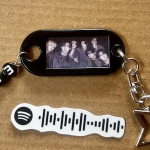 Puede incluir: Un llavero ovalado negro con una fotografía rectangular de siete personas. Incluye un dije blanco ondulado con el icono de código de Spotify y un patrón de ondas sonoras en negro. También cuenta con una estrella plateada y cuentas con letras negras.