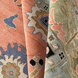 May include: Close-up of three rolled-up, intricately patterned rugs. The rugs feature a mix of colors, including coral, blue, and green, with detailed geometric and floral designs. The text "THE ANCIENT CARPETS" is visible on one of the rugs.
