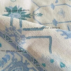 May include: Close-up of a rolled-up, off-white rug with a blue floral pattern. The rug has a textured surface and the text "THE ANCIENT CARPETS" is visible in several places.