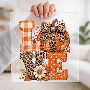 May include: A square decorative sign featuring the word LOVE in orange and brown tones. The L is orange plaid, the O is a pumpkin with a leopard print bow, the V is leopard print, and the E is orange with white polka dots and floral accents.