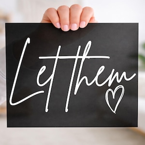 May include: A black rectangular sign with the words "Let them" in white cursive script and a white heart symbol. The sign is held up by a person with a blurred background.