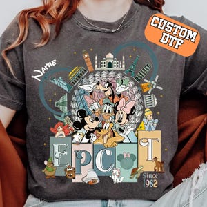 May include: Dark gray t-shirt featuring a colorful graphic of Disney characters and landmarks. The design includes Mickey and Minnie Mouse, Donald Duck, and Goofy, with the word "EPCOT" and the text "Since 1982". The shirt has the text "Name" on it and the words "Custom DTF" on an orange rectangle.