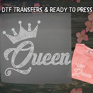 May include: A gray transfer sheet with a rhinestone crown and the word "Queen" in a script font. A pink t-shirt with a matching design is displayed next to the transfer. The text "DTF TRANSFERS & READY TO PRESS" is at the top.