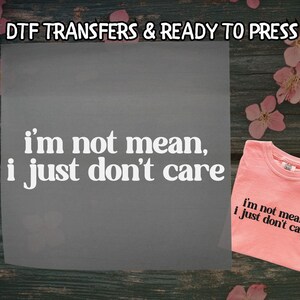 May include: A gray transfer sheet and a coral-colored t-shirt with the phrase "I'm not mean, I just don't care" printed in white. The text "DTF TRANSFERS & READY TO PRESS" is at the top. The shirt is on a wooden surface.