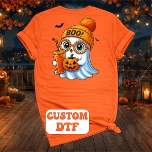 May include: An orange t-shirt featuring a graphic of a white ghost wearing an orange beanie with the text BOO!, orange round glasses, and holding an orange drink and a small pumpkin. A white rectangular label with the text CUSTOM DTF is below.