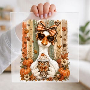 May include: A rectangular print featuring a white goose wearing heart-shaped sunglasses and a plaid leopard-print bow. The goose holds a cup with a leopard-print sleeve, surrounded by orange pumpkins, autumn leaves, and a vertical striped background.