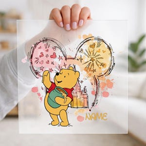 Peut inclure: Panneau acrylique transparent avec une illustration de Winnie l'Ourson tenant un pot de miel. Le dessin comprend un château, des feux d'artifice et le mot "NAME" en or. Le fond présente des éclaboussures d'aquarelle roses et jaunes.