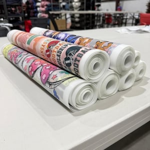 May include: Rolled fabric rolls with various designs, including sports team logos and cartoon graphics. The rolls are stacked on a white surface, showcasing the printed patterns in vibrant colors. The designs include the text "Kansas City Football" and "Philadelphia Football".