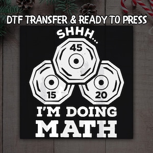 May include: Black square graphic with white text and barbell weights. The text reads "DTF TRANSFER & READY TO PRESS", "SHHH...", "45", "15", "20", and "I'M DOING MATH". The weights are white with black centers.