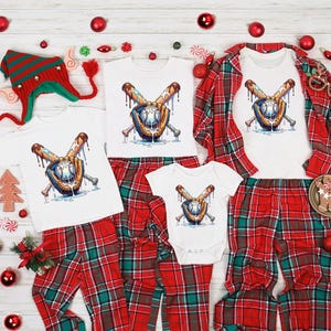 May include: A family set of matching Christmas pajamas. The set includes white t-shirts and a onesie, all featuring a baseball-themed graphic. The pajamas are paired with red and green plaid pants. Festive decorations surround the set.