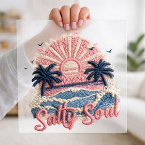 May include: Embroidered artwork featuring a sunset over the ocean with palm trees and the words "Salty Soul." The design uses pink, blue, and cream-colored thread. Small birds are also embroidered in the sky.