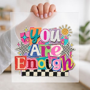 May include: A colorful graphic print with the words "YOU ARE ENOUGH" in a collage style. The design features various colors, including teal, pink, purple, and yellow, with star and sun accents. The background is clear.