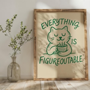May include: A framed print with a green cat illustration holding a coffee cup, with the text "EVERYTHING IS FIGUREOUTABLE". The frame is made of wood. A glass vase with greenery is to the left.
