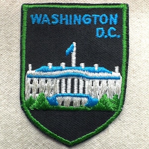 May include: Embroidered patch featuring the White House with blue accents and the text "WASHINGTON D.C." in blue. The patch has a black background with a green border, showcasing a shield shape.