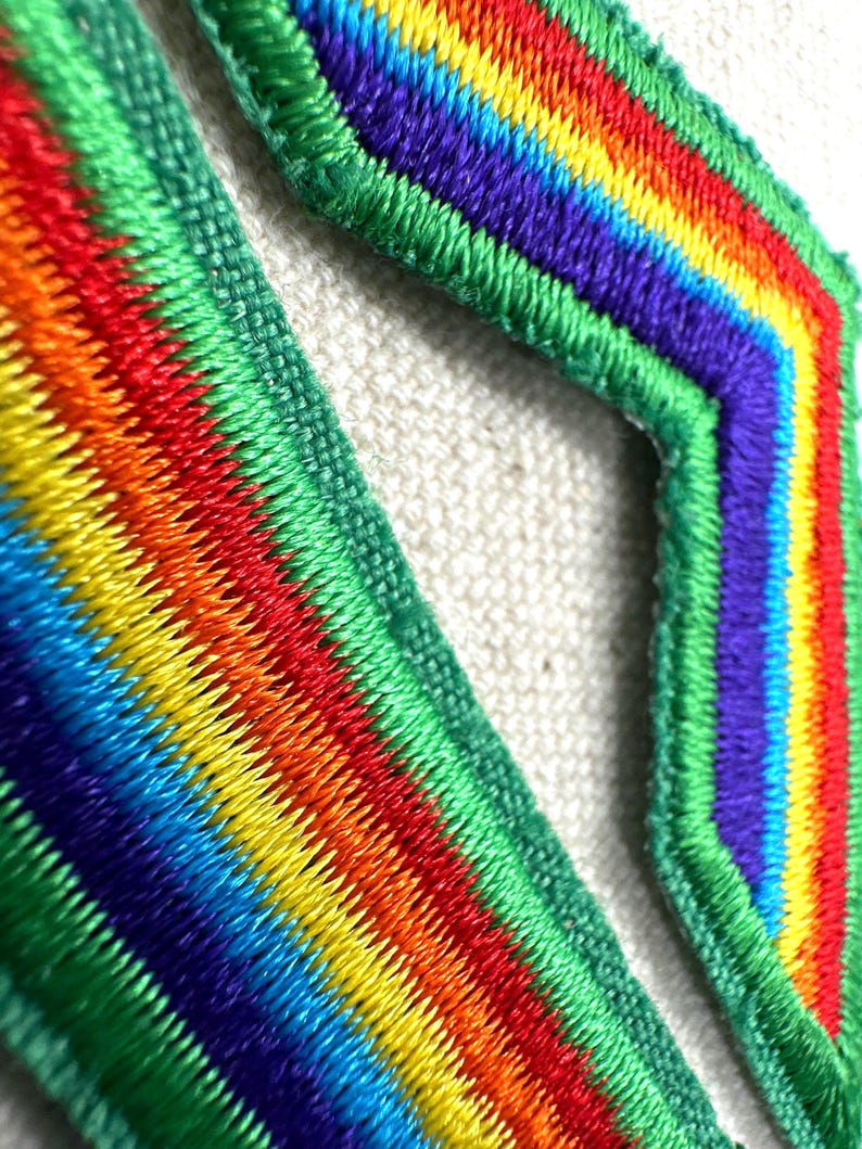 Puede incluir: Primer plano de dos parches bordados con un dise&ntilde;o de arco&iacute;ris. Los parches presentan colores vibrantes que incluyen rojo, naranja, amarillo, verde, azul y morado. Los bordes est&aacute;n delineados con hilo verde.