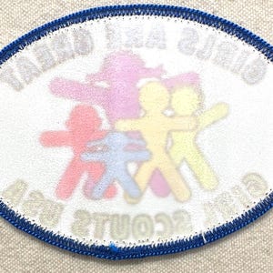 May include: Oval patch with a blue border and white background. The patch features colourful stick figures in red, purple, blue, and yellow. The text "GIRLS AND GREAT SCOUTS" is visible.