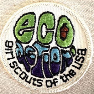 May include: Circular embroidered patch with the words "Eco Action" in green, blue, and purple, with the text "Girl Scouts of the USA" in black. The patch has a white border.