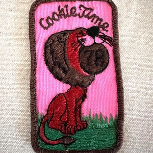 Puede incluir: Parche bordado con las palabras "Cookie Time" sobre un le&oacute;n de dibujos animados que sostiene una galleta. El parche tiene un fondo rosa, un borde marr&oacute;n y hierba verde en la parte inferior. El le&oacute;n es rojo y marr&oacute;n.