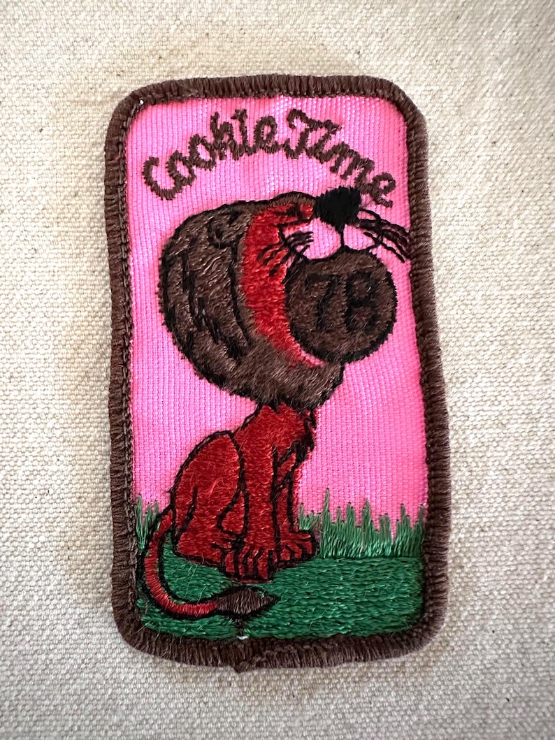 Puede incluir: Parche bordado con las palabras "Cookie Time" sobre un le&oacute;n de dibujos animados que sostiene una galleta. El parche tiene un fondo rosa, un borde marr&oacute;n y hierba verde en la parte inferior. El le&oacute;n es rojo y marr&oacute;n.