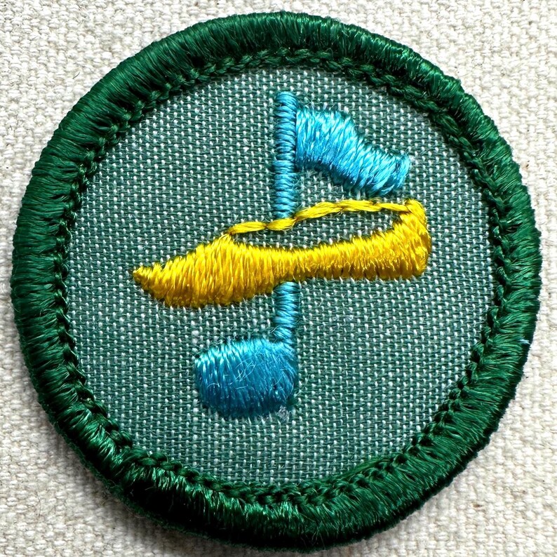 May include: Embroidered patch with a circular design. The patch features a teal background with a green border. The design includes a blue and yellow abstract shape. The patch is likely for a uniform or craft project.