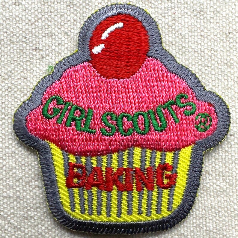 Puede incluir: Parche de cupcake bordado con una cereza roja. El glaseado rosa tiene las palabras "GIRL SCOUTS" en verde. La base amarilla de la magdalena tiene la palabra "BAKING" en rojo. El parche est&aacute; delineado en gris.