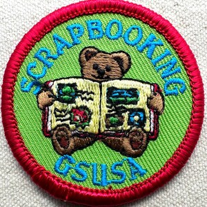 May include: Embroidered patch with a green background and a red border. The patch features a brown teddy bear holding a scrapbook. The words "SCRAPBOOKING" and "GSUSA" are embroidered in blue.