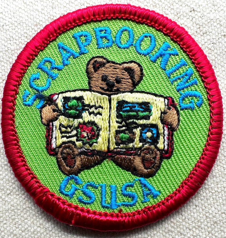 May include: Embroidered patch with a green background and a red border. The patch features a brown teddy bear holding a scrapbook. The words "SCRAPBOOKING" and "GSUSA" are embroidered in blue.