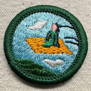 May include: Embroidered patch featuring a person in green clothing on a yellow rug, floating over a blue background with white clouds. The patch has a green border and is likely for a scout or similar organization.