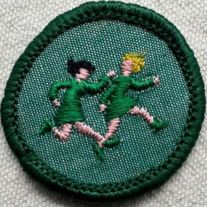Puede incluir: Un parche redondo bordado en verde con dos figuras con vestidos verdes corriendo. Las figuras tienen piernas y brazos rosas, una con cabello negro y la otra rubio. El parche tiene un borde verde.